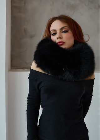 A woman with red hair wearing a black off-the-shoulder knit sweater and a voluminous black fur collar, posed against a textured gray wall.